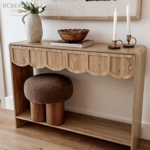 May include: A light oak console table with a scalloped front edge and a lower shelf. A brown textured round stool sits underneath. On top are a white ceramic vase with white tulips, a wooden bowl on a book, and two white candles in brass holders.