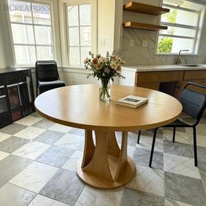 May include: A round light oak dining table with a central pedestal base featuring curved supports. A clear glass vase filled with a bouquet of white, pink, and yellow flowers sits in the center of the table next to a small white rectangular book.