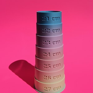 Puede incluir: Una pila de tazas medidoras graduadas, cada una con una medida en centímetros de 21 cm a 27 cm. Las tazas varían en color de azul a rosa, sobre un fondo rosa brillante.