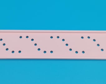 May include: A light pink wooden ruler with a series of evenly spaced holes arranged in a wavy pattern. The ruler is rectangular and has a raised edge on both sides. The background is a solid blue.