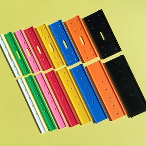 Può includere: Una collezione di strisce di plastica colorate in bianco, verde, rosa, rosso, giallo, blu, arancione e nero. Ogni striscia ha più fori e una fessura centrale. Gli oggetti sono disposti su una superficie gialla.