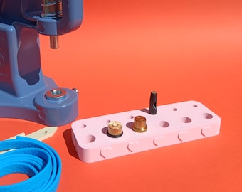 May include: A blue press machine with a silver spring and a pink tray with various metal tools. A blue strip of material and a white tool are also present. The background is a solid orange color.