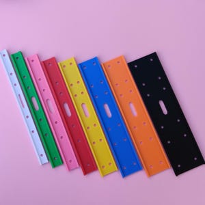 Puede incluir: Una colección de tiras de plástico rectangulares de colores blanco, verde, rosa, rojo, amarillo, azul, naranja y negro. Cada tira tiene múltiples agujeros y una abertura central alargada. Las tiras están dispuestas en diagonal sobre un fondo rosa.