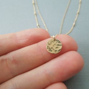 May include: A delicate gold necklace with a hammered circle pendant. The necklace is layered with two chains, one with tiny beads and the other a delicate chain.