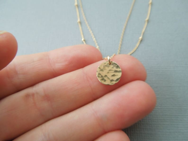 May include: A delicate gold necklace with a hammered circle pendant. The necklace is layered with two chains, one with tiny beads and the other a delicate chain.