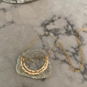 May include: A gold necklace with a hammered double crescent moon pendant. The necklace is on a white marble surface.