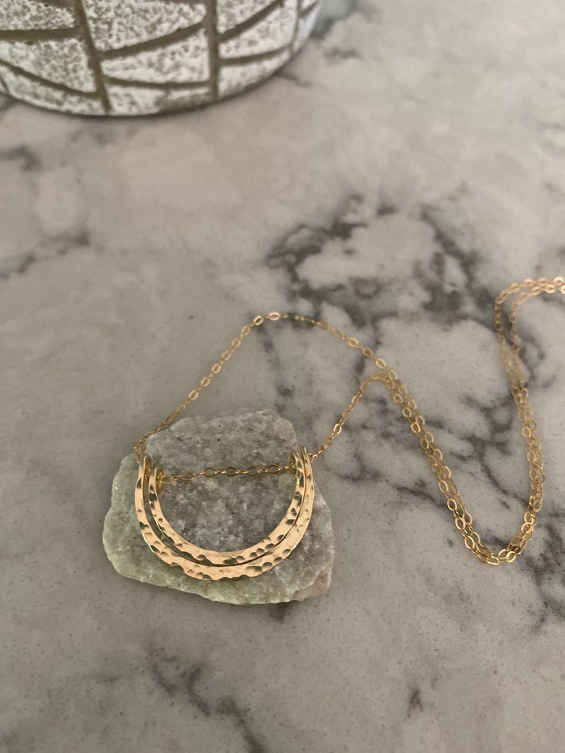 May include: A gold necklace with a hammered double crescent moon pendant. The necklace is on a white marble surface.