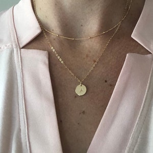 May include: A gold chain necklace with a small round pendant featuring the letter 'A'. The necklace is layered over a thinner gold chain.
