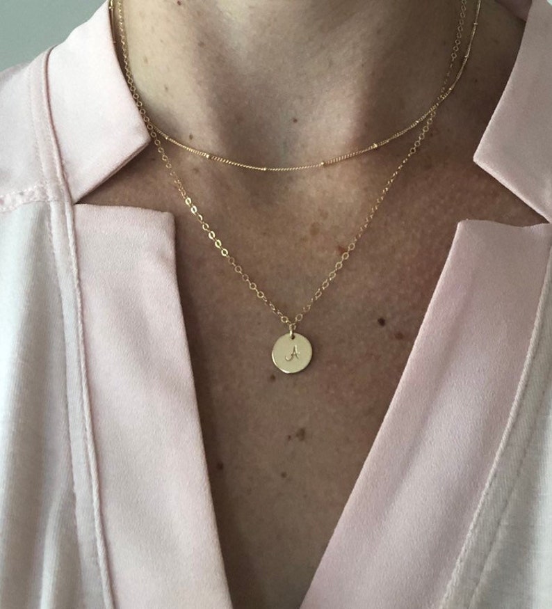 May include: A gold chain necklace with a small round pendant featuring the letter 'A'. The necklace is layered over a thinner gold chain.