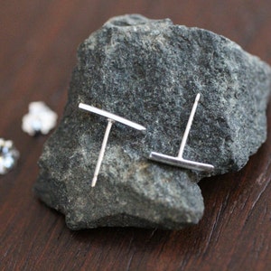 May include: A pair of silver stud earrings with a horizontal bar design. The earrings are displayed on a gray rock.