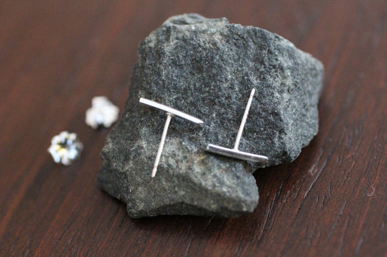 May include: A pair of silver stud earrings with a horizontal bar design. The earrings are displayed on a gray rock.