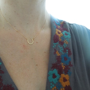 Peut inclure: Un collier en forme de fer &agrave; cheval en or port&eacute; par un mannequin v&ecirc;tu d'une chemise grise avec des fleurs brod&eacute;es.