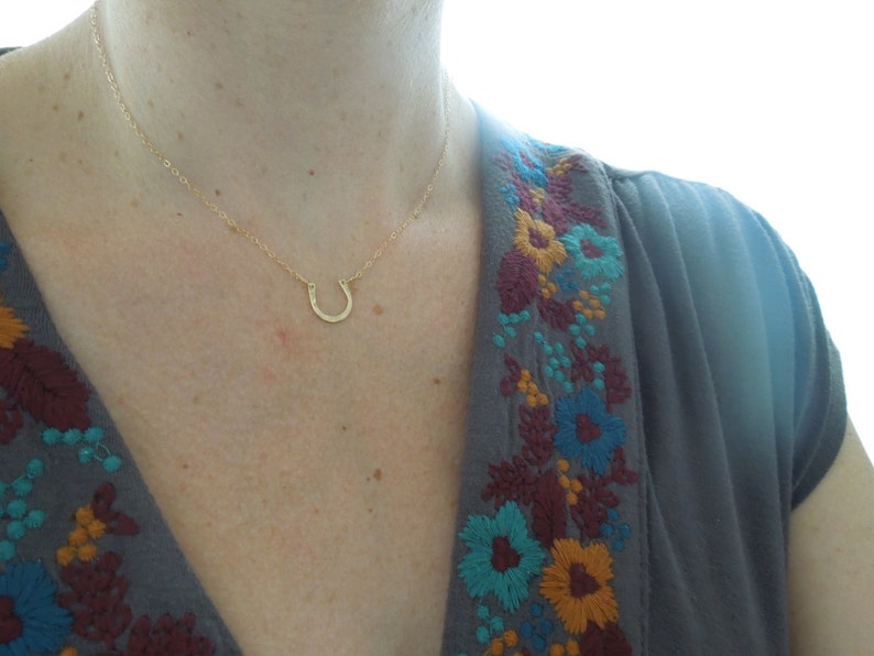 Peut inclure: Un collier en forme de fer &agrave; cheval en or port&eacute; par un mannequin v&ecirc;tu d'une chemise grise avec des fleurs brod&eacute;es.