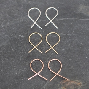 May include: Three pairs of simple, minimalist earrings in silver, gold, and rose gold. Each pair features a thin, looped wire that crosses over itself to form a delicate, modern design.