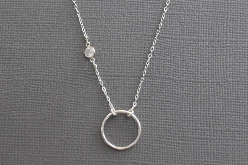 Puede incluir: Un collar de cadena de plata con un peque&ntilde;o acento de piedra clara y un colgante circular simple.
