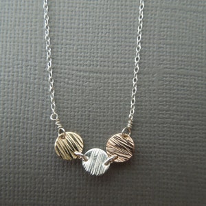 May include: A silver chain necklace with three small, round pendants. The pendants are made of different metals: gold, silver, and rose gold. Each pendant has a textured surface.