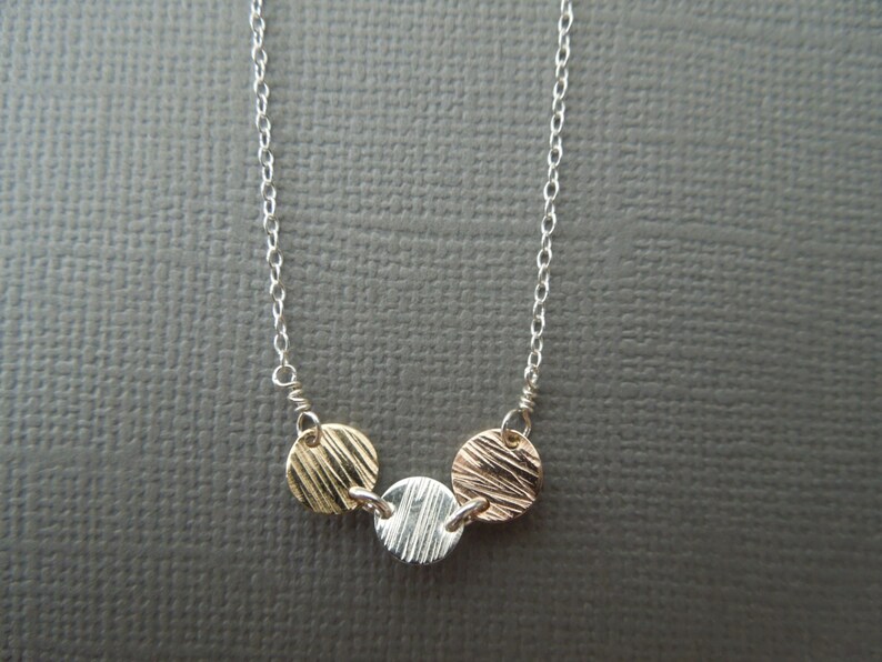 May include: A silver chain necklace with three small, round pendants. The pendants are made of different metals: gold, silver, and rose gold. Each pendant has a textured surface.