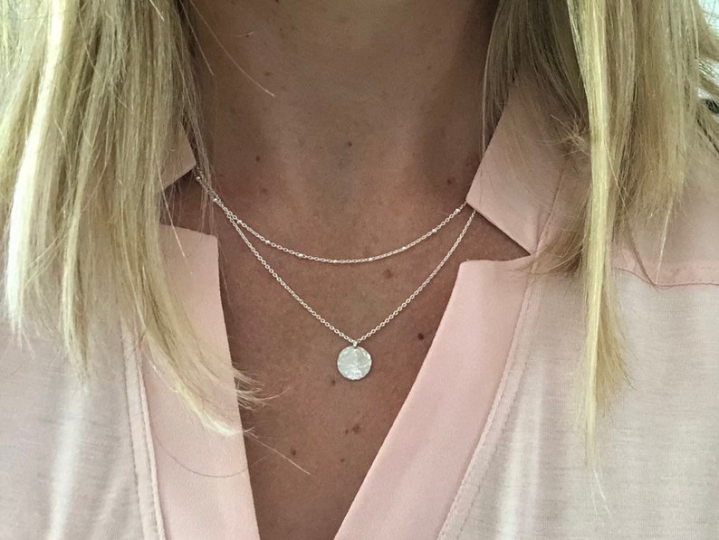 May include: A silver necklace with a delicate chain and a small, round pendant. The pendant has a hammered texture and is a simple, minimalist design.