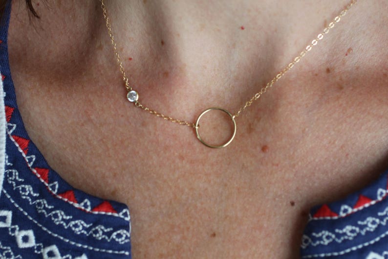 Puede incluir: Un collar de cadena de oro con un peque&ntilde;o colgante circular y un solo acento de piedra clara.