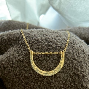 May include: A gold necklace with a hammered double arch pendant. The necklace is on a brown fuzzy surface.