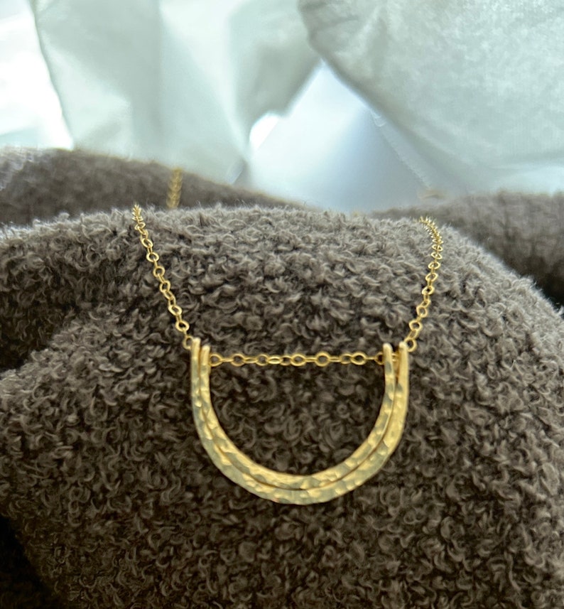 May include: A gold necklace with a hammered double arch pendant. The necklace is on a brown fuzzy surface.