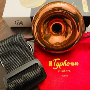 May include: A copper-colored mini horn with a black strap and a red pouch. The pouch has the text "Typhoon minihorn TAIWAN" printed on it.