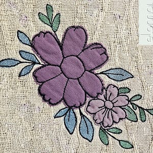 Könnte beinhalten: Gestickte lila und blaue Blumen mit grünen Blättern auf einem beigefarbenen Stoffhintergrund.