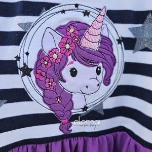 May include: A white and navy blue striped shirt with a purple unicorn embroidered on the front. The unicorn has a pink horn, a purple mane, and a pink flower crown. The shirt has a purple ruffle at the bottom.