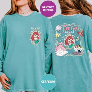 a woman wearing a teal long-sleeved shirt with a design of Ariel from the Disney movie "The Little Mermaid". The design includes Ariel's face, a mermaid, and various other characters from the film. The shirt also has a "Next day shipping" label, indicating that the shirt is available for immediate purchase.