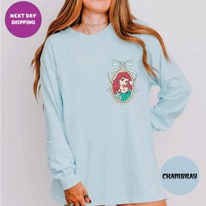 A woman wearing a light blue long-sleeved shirt with a graphic of a mermaid on the front.