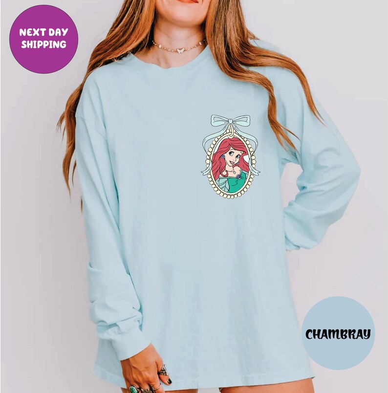 A woman wearing a light blue long-sleeved shirt with a graphic of a mermaid on the front.