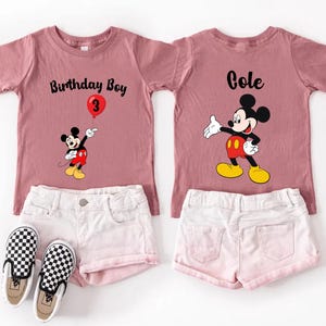 a pink t-shirt with a Mickey Mouse design and the text "Birthday Boy 3" printed on it, along with a pair of white shorts and checkered shoes.