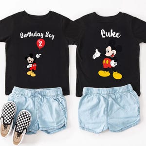 a pair of black t-shirts with Mickey Mouse designs, paired with denim shorts and checkered shoes, suggesting a birthday outfit for a child.
