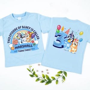 May include: Two light blue t-shirts with a colorful graphic featuring characters from the animated series Bluey. One shirt says "This episode of Bluey is called Marshall turns three!" The other shirt has a large number 3 with balloons and confetti.