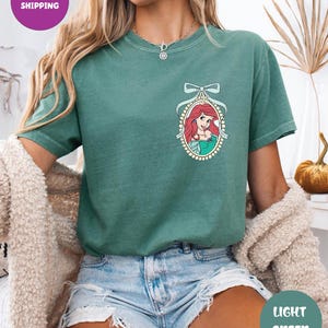 A woman wearing a green t-shirt with a mermaid graphic and denim shorts, sitting in a room with a beige sweater and a plant in the background.