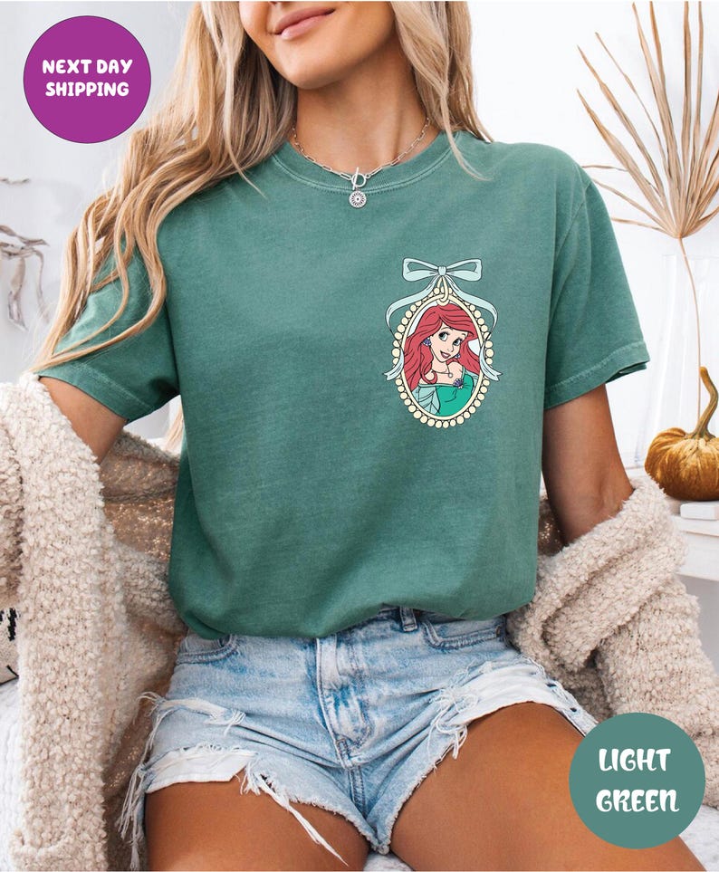 A woman wearing a green t-shirt with a mermaid graphic and denim shorts, sitting in a room with a beige sweater and a plant in the background.