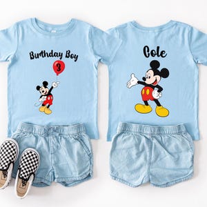 two blue t-shirts with Mickey Mouse designs, each with a personalized text and a balloon graphic, along with matching denim shorts and black and white checkered shoes.