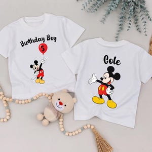 two white t-shirts with Mickey Mouse designs, one labeled "Birthday Boy" and the other labeled "Cole", along with a teddy bear and some beads.