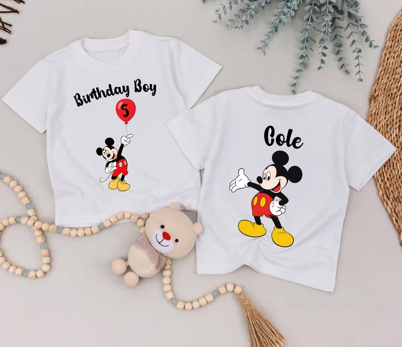 two white t-shirts with Mickey Mouse designs, one labeled "Birthday Boy" and the other labeled "Cole", along with a teddy bear and some beads.
