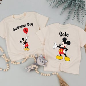 two white t-shirts with Mickey Mouse designs, one labeled "Birthday Boy" and the other labeled "Cole", along with a cat toy and some greenery.