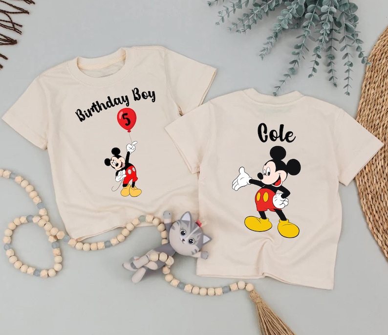 two white t-shirts with Mickey Mouse designs, one labeled "Birthday Boy" and the other labeled "Cole", along with a cat toy and some greenery.
