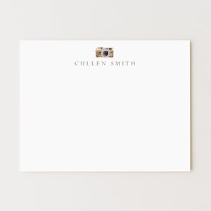 May include: A white stationery card with a watercolor illustration of a vintage camera at the top. The name "CULLEN SMITH" is printed in a dark gray font below the camera image. The card is rectangular.