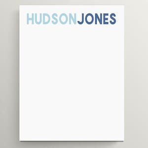 May include: A white notepad with the name "HUDSON JONES" printed in shades of blue across the top. The notepad is rectangular and has a clean, minimalist design, suitable for notes or lists.