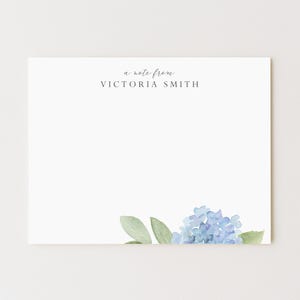 May include: White stationery with the text "a note from VICTORIA SMITH" in a delicate font. A watercolor illustration of blue hydrangea flowers and green leaves adorns the bottom right corner. Stationery for personal use.