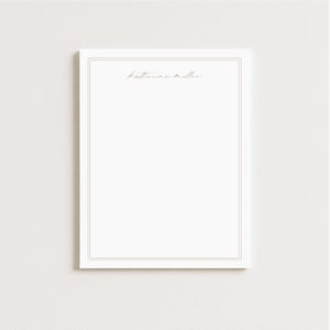 May include: A white notepad with a double border and the name "Katherine Miller" in a delicate script at the top. The notepad is rectangular and sits against a plain, off-white background.