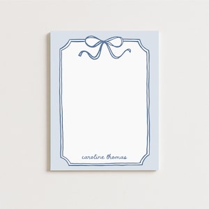 May include: A light blue notepad with a dark blue border and a bow design at the top. The name "caroline thomas" is written in dark blue at the bottom of the notepad.