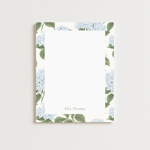 May include: A rectangular notepad with a white interior and a floral border. The border features light blue hydrangea blooms and green leaves against a cream background. The name "Ella Thomas" is printed in a small font at the bottom.