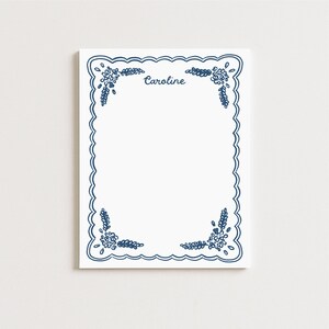 May include: A white notepad with a scalloped blue border and floral corner designs. The name "Caroline" is written in blue script at the top. The notepad is rectangular and has a clean, minimalist design.