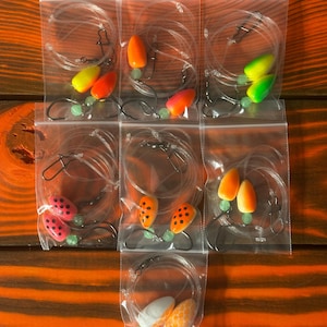 May include: Assortment of fishing lures in clear plastic bags. The lures feature various color combinations, including orange, yellow, red, and white, with black spots on some. Each lure is attached to a fishing line with a hook and a small green bead.