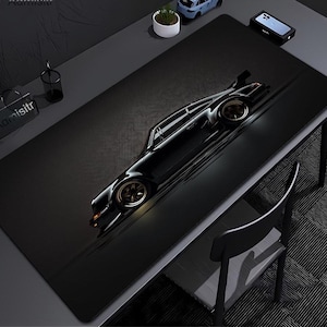 May include: A large rectangular black desk mat featuring a high-contrast graphic of a sleek black sports car in profile. The car has gold-toned wheels and is positioned diagonally across the center of the mat, which has a subtle textured surface.